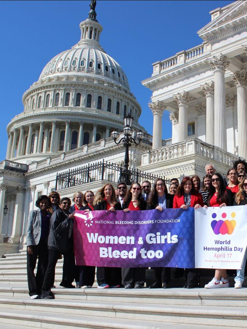 Women and girls with bleeding disorders standing on the steps of the capitol building in Washington DC, holding a banner that says "Women and girls bleed too"