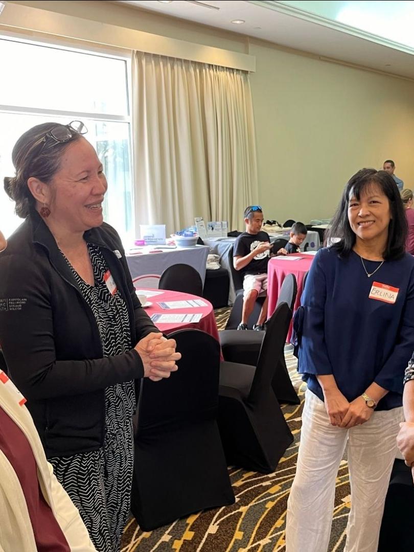 Four women are standing in a circle, talking and smiling at each other at a community event.. In the background, there are round tables with handouts and a few other people talking with each other.