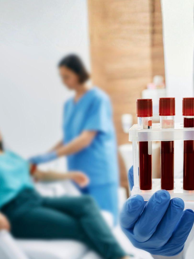 A series of blood vials are in the foreground, being held by a pair of blue-gloved hands. Blurred in the background is a woman lying in on an examination table, being attended to by a woman in blue scrubs.