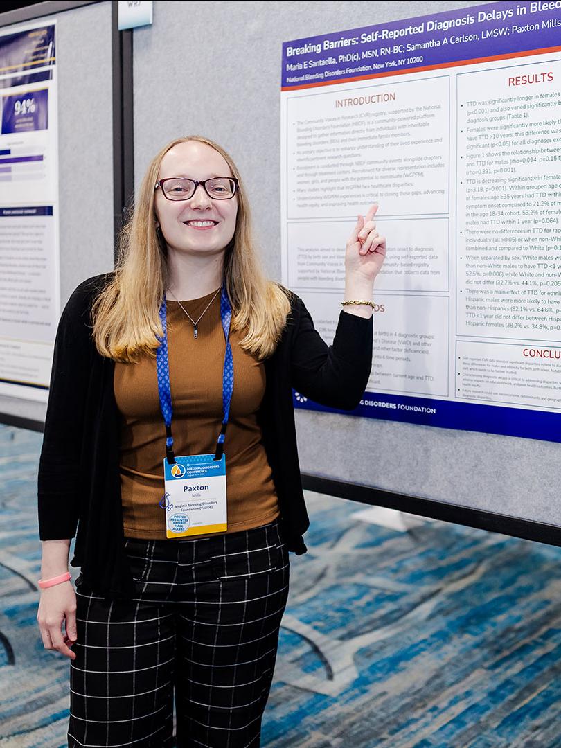A young woman, Paxton Mills, stands in front of a research poster abstract, pointing at the data