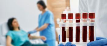 A series of blood vials are in the foreground, being held by a pair of blue-gloved hands. Blurred in the background is a woman lying in on an examination table, being attended to by a woman in blue scrubs.