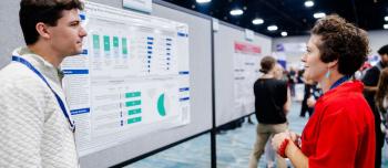 A man and a woman are standing in front of a research poster abstract in a large exhibit hall